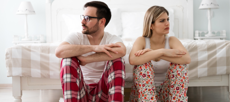 a man and a woman in pajamas sit next to each other, looking away from one another and clearly upset