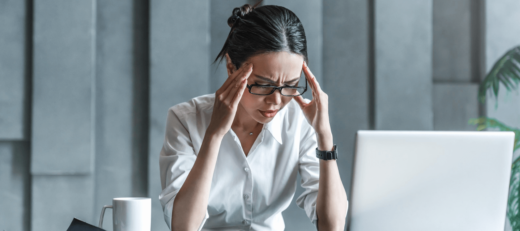 a woman with glasses in pain holds her head while looking at a laptop